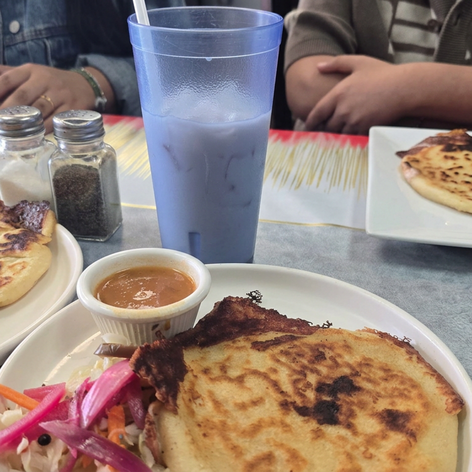 Table set with grilled flatbreads, dipping sauce, pickled vegetables, spices, and a blue drink, with diners seated nearby.